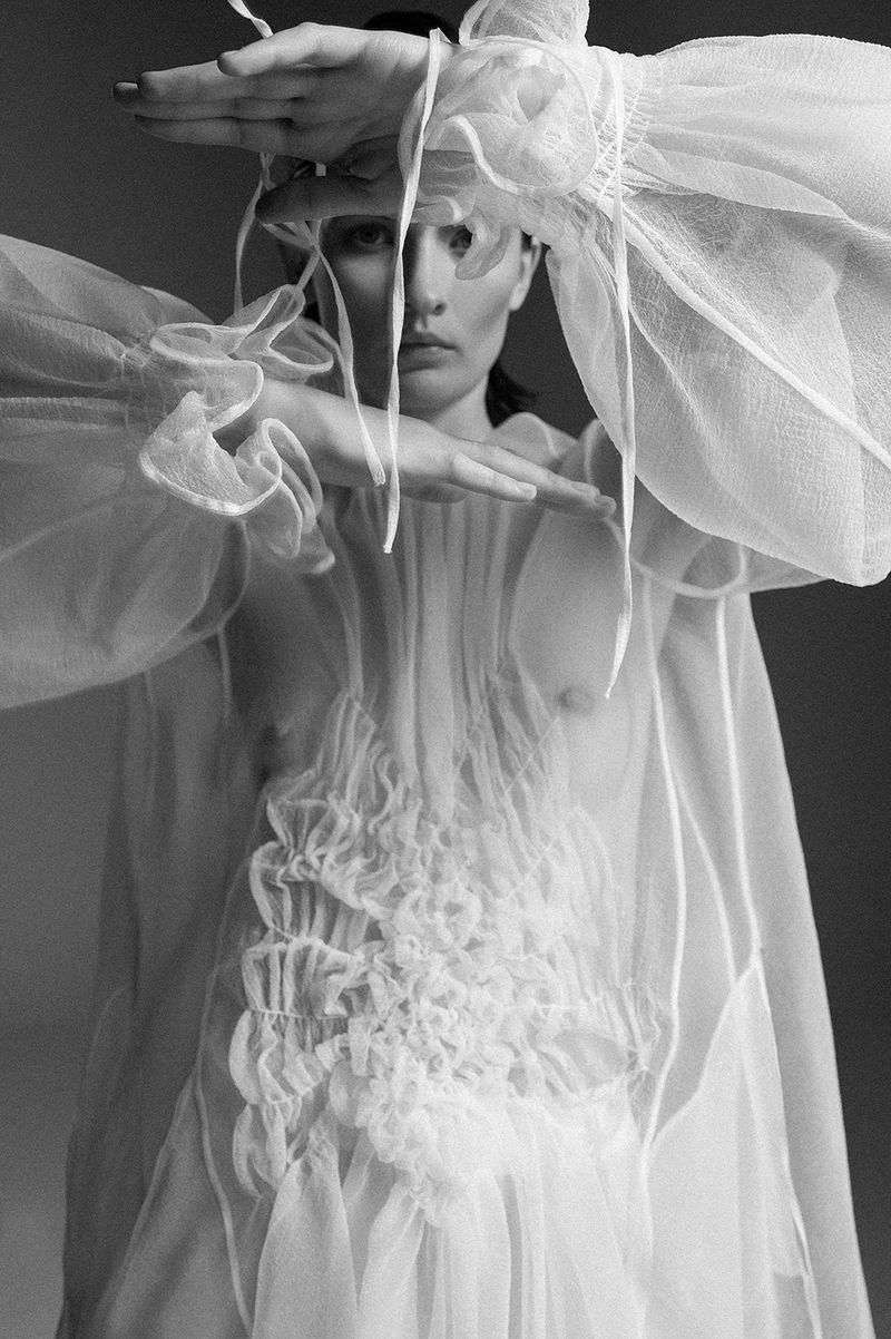 Black and white portrait of a woman in a sheer white gown, framing her face with her hands in a photo studio setting.