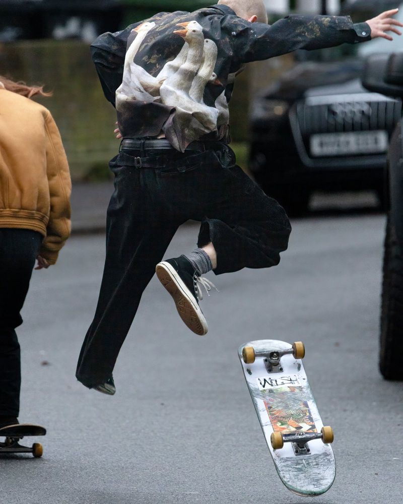 Young person mid-air on a skateboard, performs a trick on the street with another skateboarder and cars in the background.