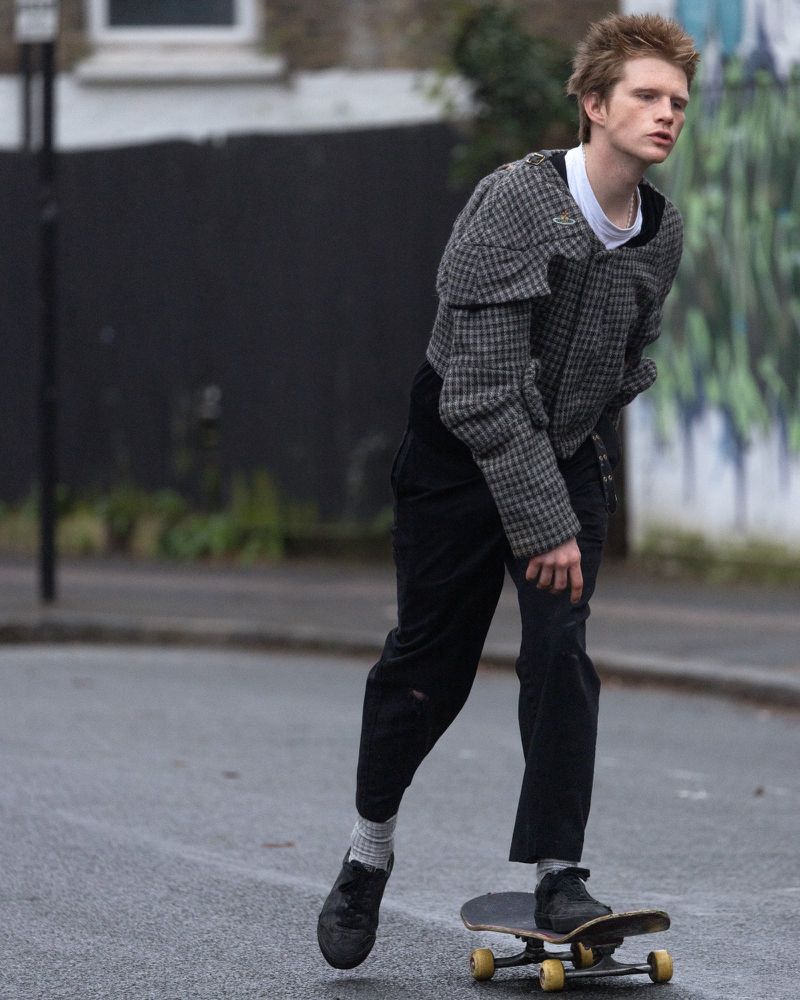 A young adult male with reddish blond hair rides a skateboard in an urban setting, wearing a patterned jacket and black pants on a cloudy day.