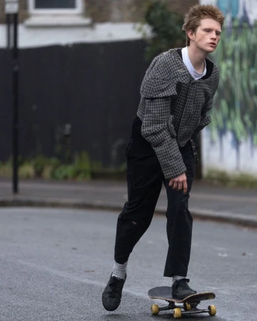 A young adult male with reddish blond hair rides a skateboard in an urban setting, wearing a patterned jacket and black pants on a cloudy day.