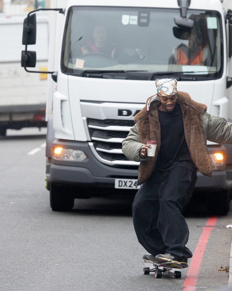 A man with head scarf skateboarding down city street, holding a coffee cup. A large white truck is behind him.