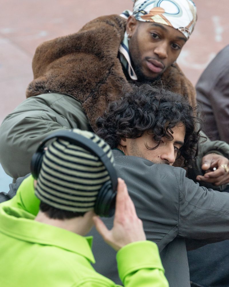 Three adult men with fashionable attire are posing outdoors. One man wears a headscarf and fur coat, while another has headphones and a green jacket.