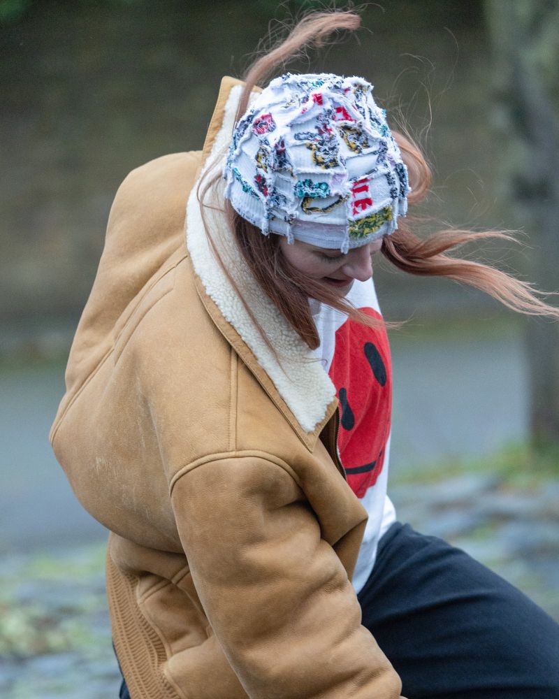 A young woman with long red hair wears a white and multicolored patchwork beanie and a tan shearling jacket against a blurred background.