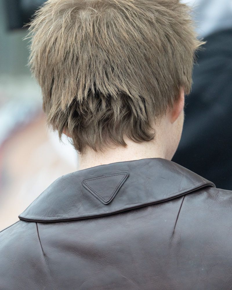 Close-up of the back of an adult with short brown hair wearing a brown leather jacket. A triangular logo is visible on the jacket collar.