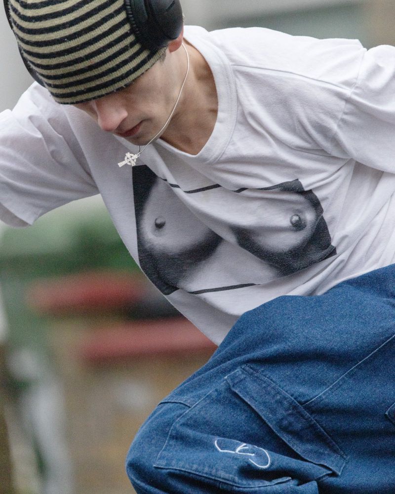 Close-up of a young adult wearing a white tee with a graphic design, a striped beanie, and large headphones in an outdoor urban setting.