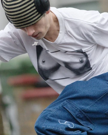 Close-up of a young adult wearing a white tee with a graphic design, a striped beanie, and large headphones in an outdoor urban setting.