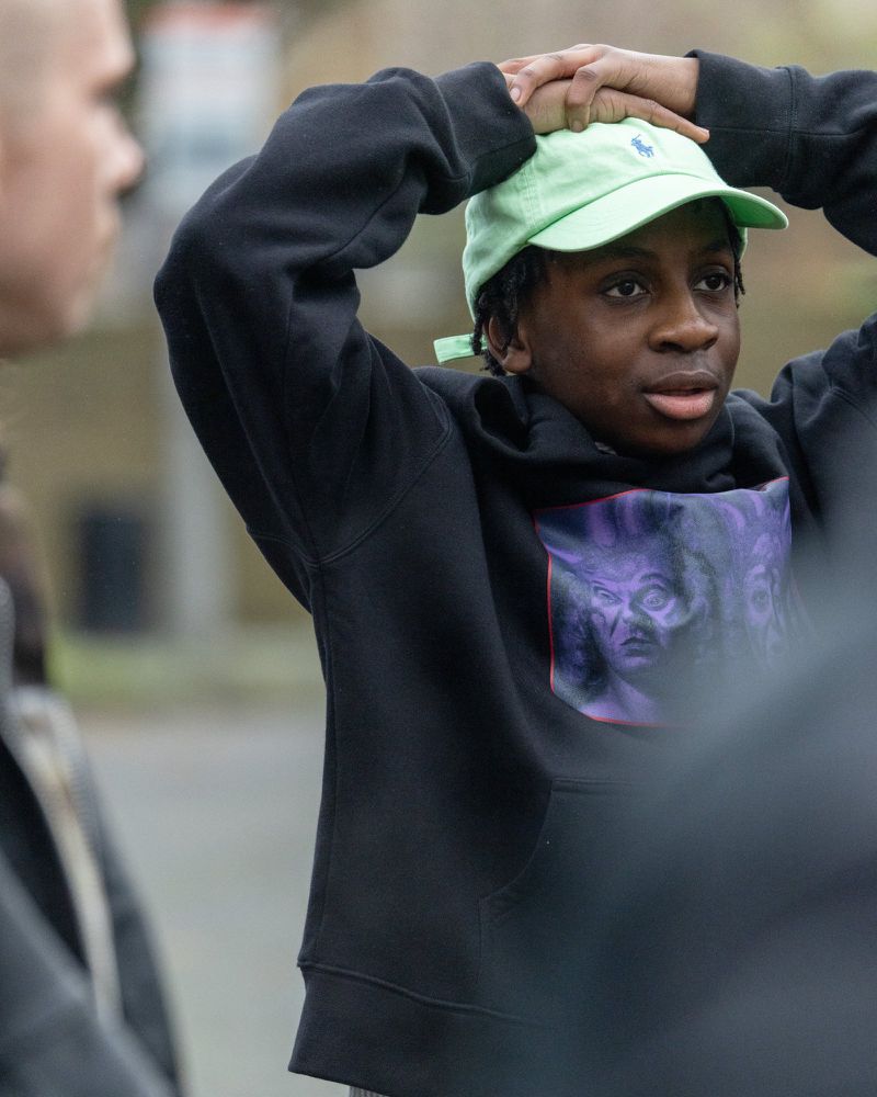 Teenage boy in a black hoodie and green baseball cap stands outside with his hands on his head looking off to the side.