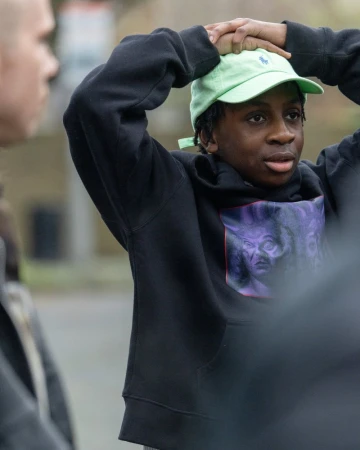 Teenage boy in a black hoodie and green baseball cap stands outside with his hands on his head looking off to the side.