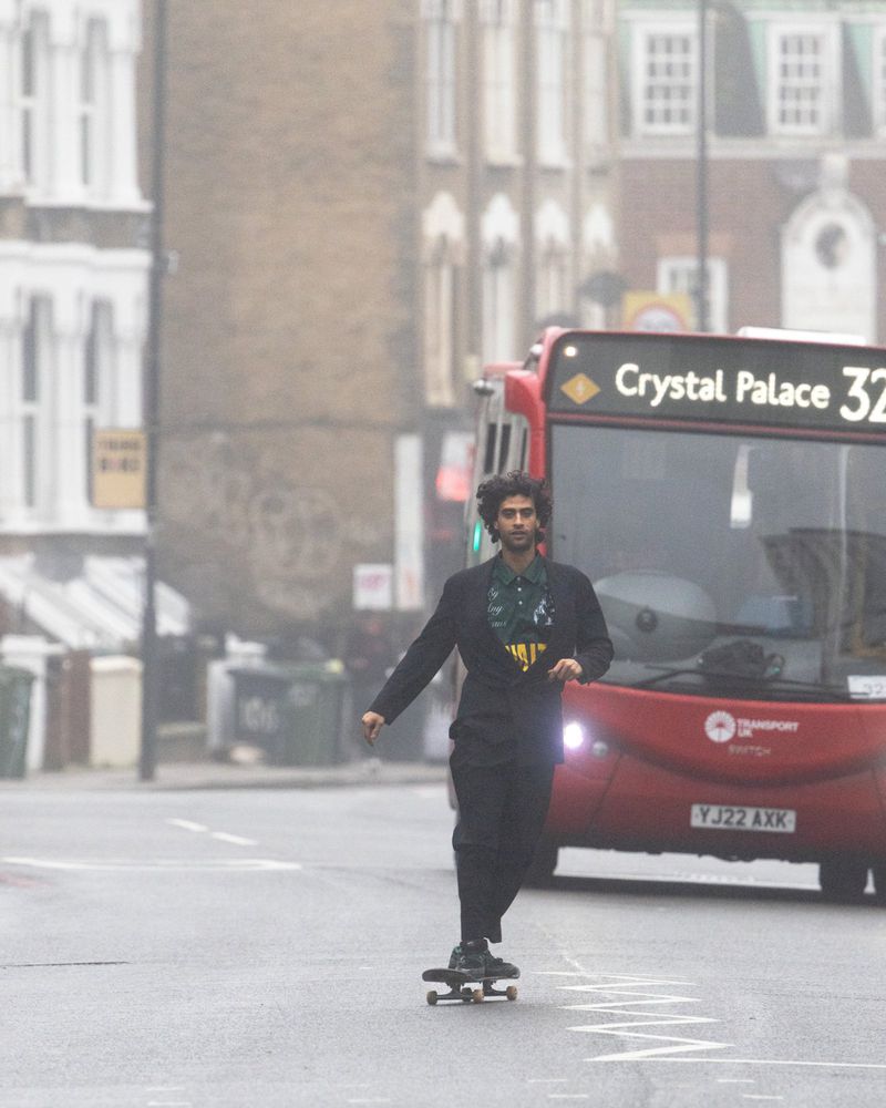 A man rides a skateboard on a city street near a double decker bus. He wears a blazer and trousers. Buildings are in the background.