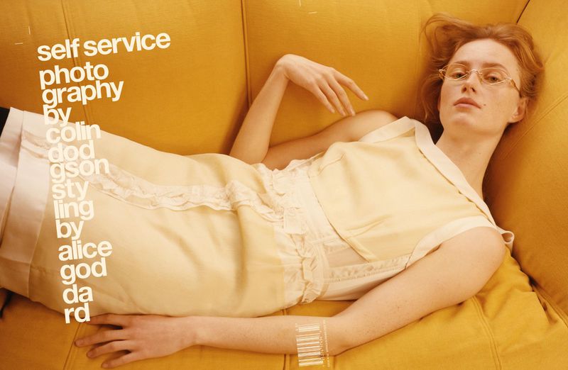 Adult female model poses on a yellow couch, wearing vintage eyeglasses and a pastel yellow dress in a fashion editorial shot.