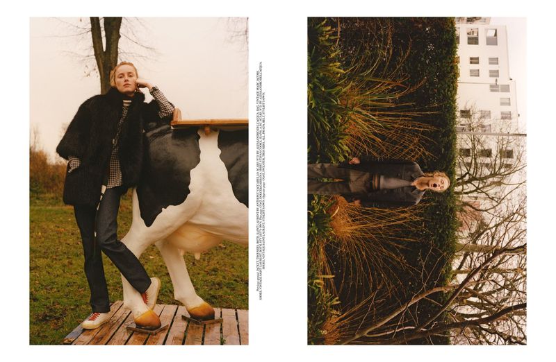Fashion editorial photo featuring a female model in a black coat posing next to a large cow sculpture in a rural outdoor setting.