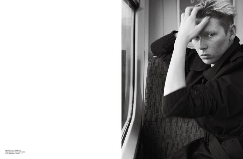 Black and white portrait of a fair-skinned adult in a dark trench coat sitting on a train, glancing at the viewer while resting their head in their hand.