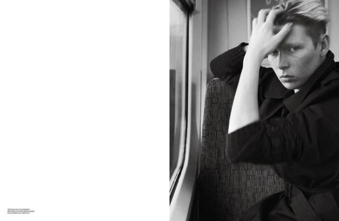 Black and white portrait of a fair-skinned adult in a dark trench coat sitting on a train, glancing at the viewer while resting their head in their hand.
