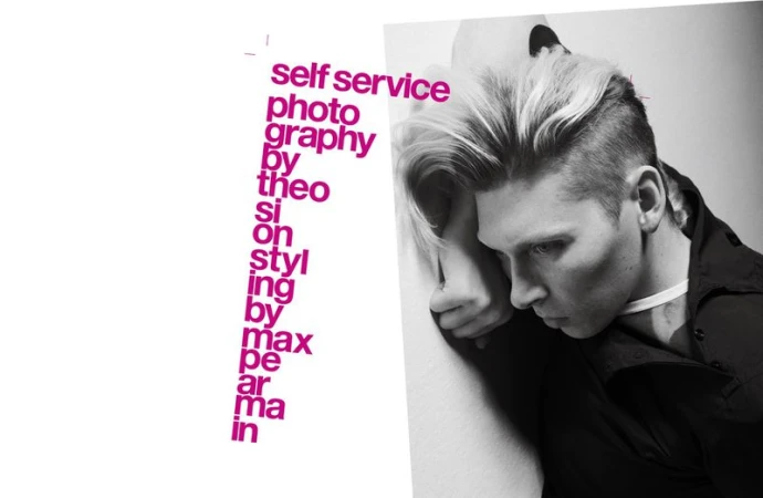 Monochrome fashion portrait of an adult male with blond hair, wearing a black shirt and white choker, posed against a light background.