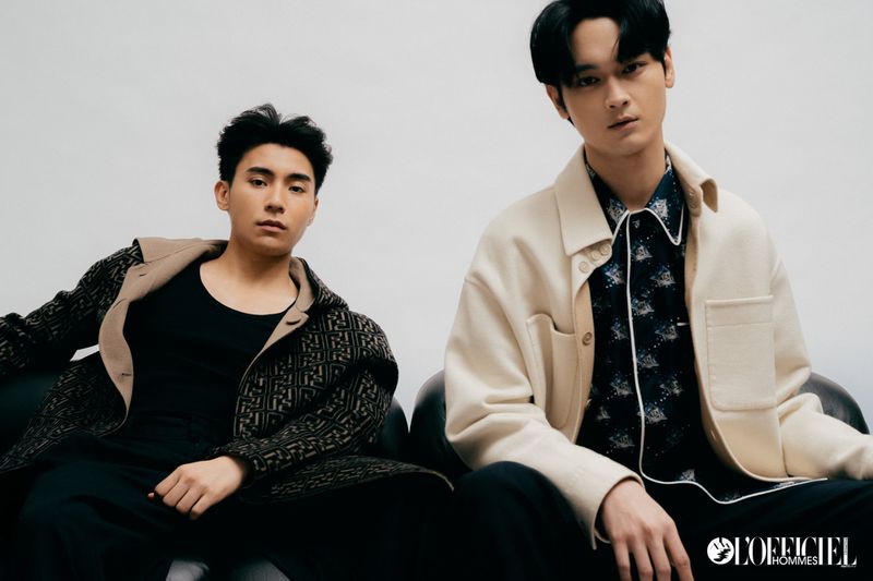 Two young Asian men pose indoors in fashionable outfits, one in a geometric jacket and the other in a shirt and cream jacket.