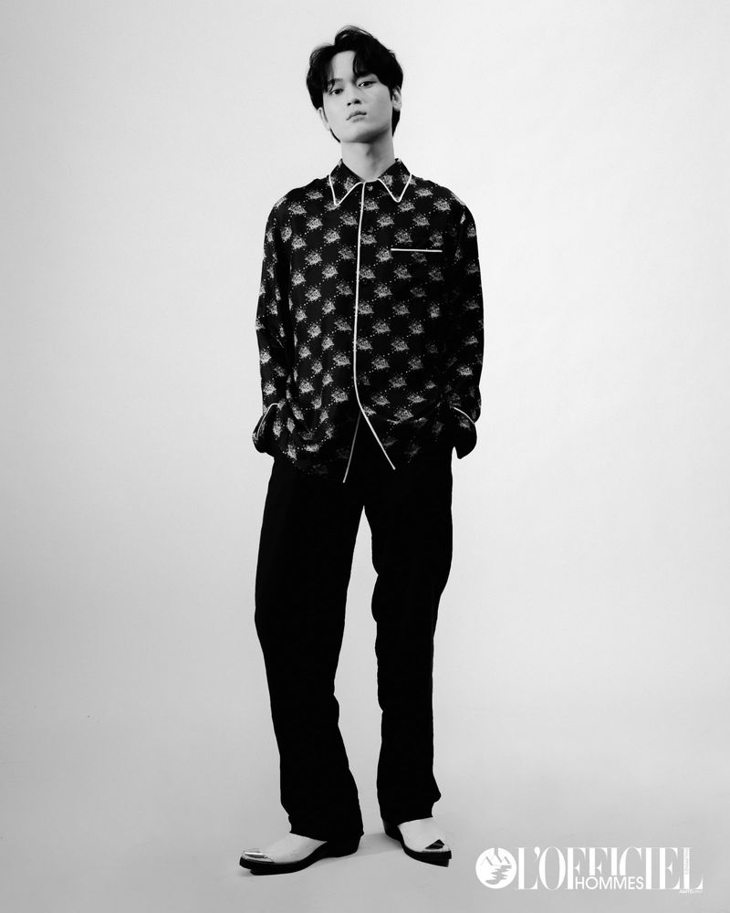 Portrait of a young adult man in a patterned button-down shirt and dark pants, posing indoors against a light background in a studio shot.