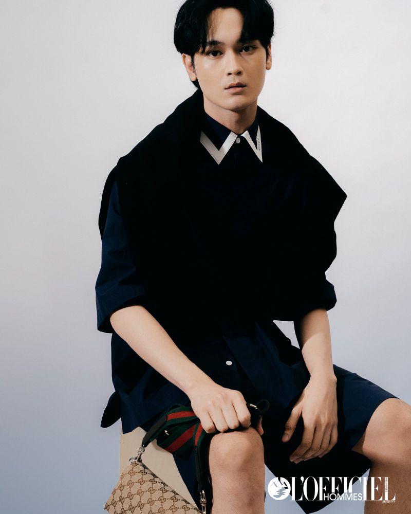 Portrait of a young Asian man wearing a navy shirt and black vest, seated and holding a Gucci handbag in a studio with soft lighting.