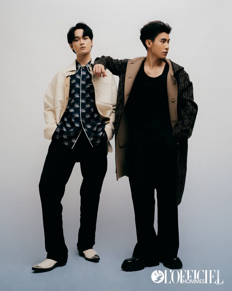 Two adult men pose indoors wearing stylish outerwear, black pants, and sleek dress shoes in a studio setting. L'Officiel Hommes logo visible.
