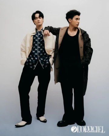 Two adult men pose indoors wearing stylish outerwear, black pants, and sleek dress shoes in a studio setting. L'Officiel Hommes logo visible.