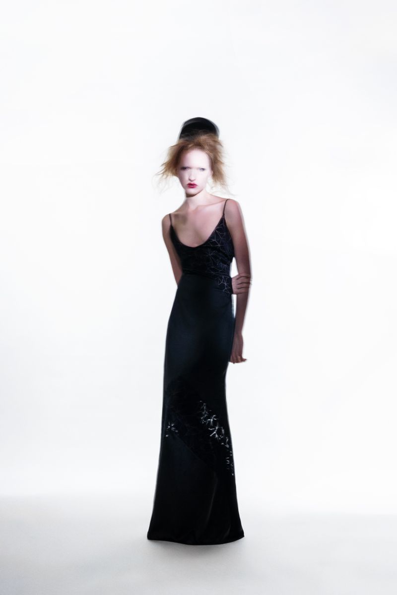 Elegant woman in a black evening gown stands in a full shot on a white background, showcasing a sophisticated and fashionable look.
