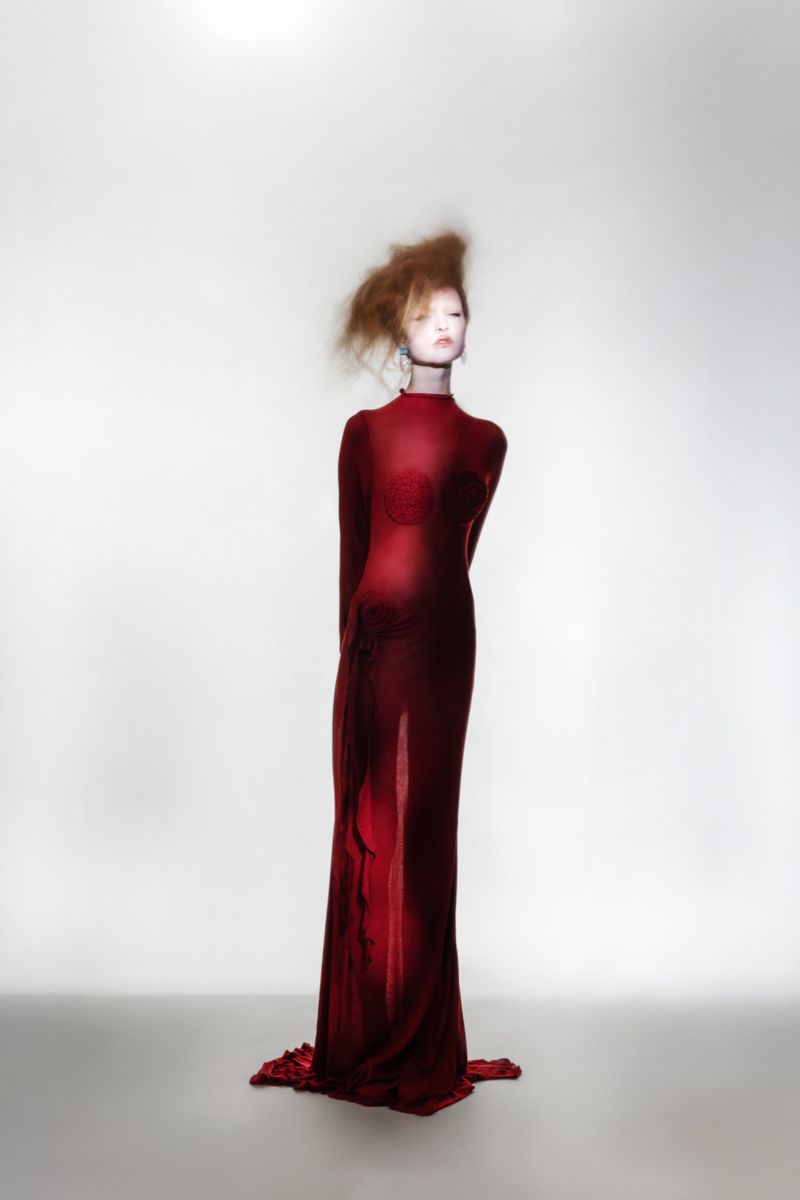 Woman in a long red gown poses in a photo studio against a light gray backdrop, wearing an elegant long dress with red hair.
