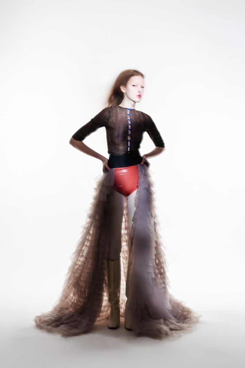 A fashion photo shows a slender woman in a unique outfit consisting of a sheer black top, orange bottoms, tan boots, and a tulle skirt.