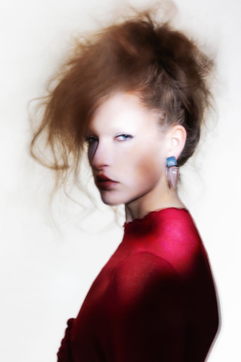 Woman with distinctive makeup and earrings, red top in a blurred photo studio setting.