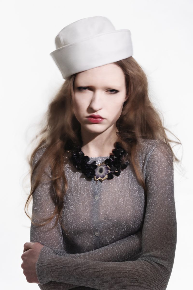 Portrait of a woman with auburn hair wearing a white pillbox hat, statement necklace, and sheer cardigan against a white backdrop.