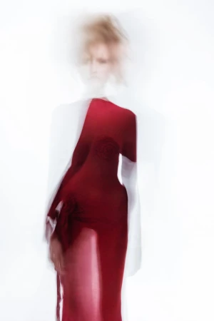 Blurry model wearing a long red dress against a bright white background in a fashion editorial shot.