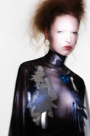 Model with reddish hair wearing a dark shiny top and blue crystal pendant against a bright white background