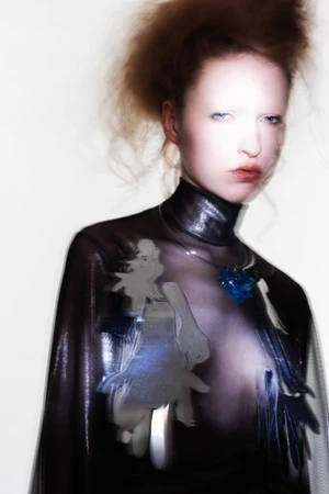 Model with reddish hair wearing a dark shiny top and blue crystal pendant against a bright white background