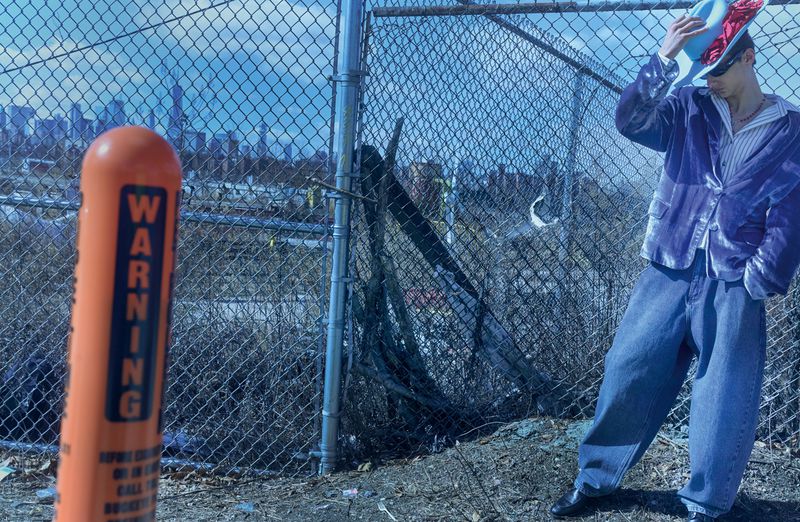 Man in blue velvet jacket and wide-leg jeans poses near a chain-link fence with an urban skyline in the background. Bright daylight.