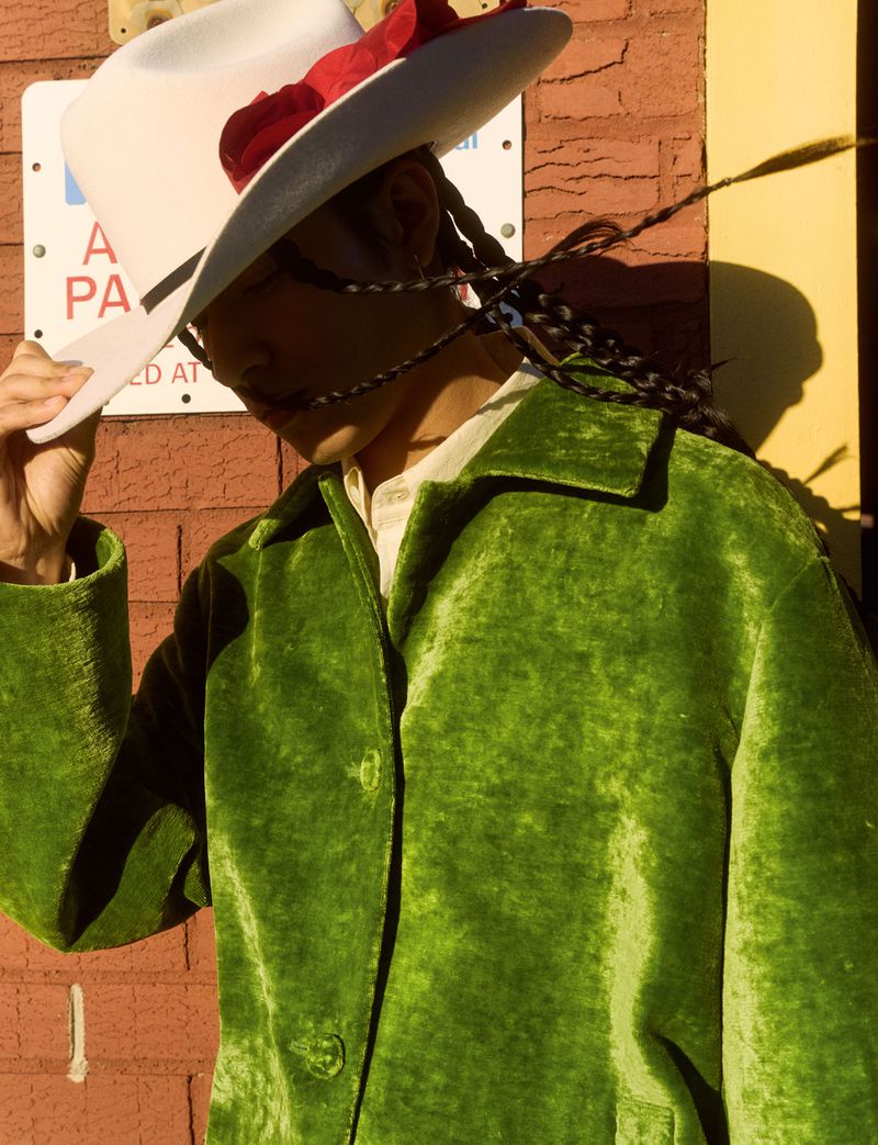 Person wearing a green velvet coat and white cowboy hat adorned with red flowers standing against a brick wall with signage.