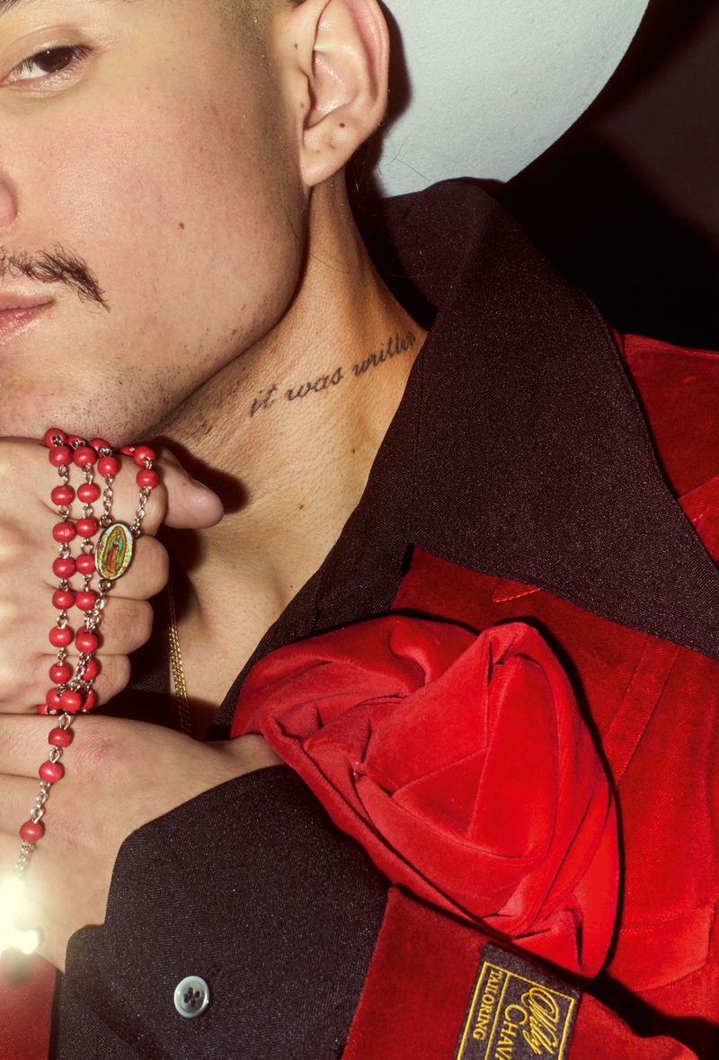 Portrait of a young adult wearing a red and black coat and a white hat, holding red beaded rosary beads against his shoulder.