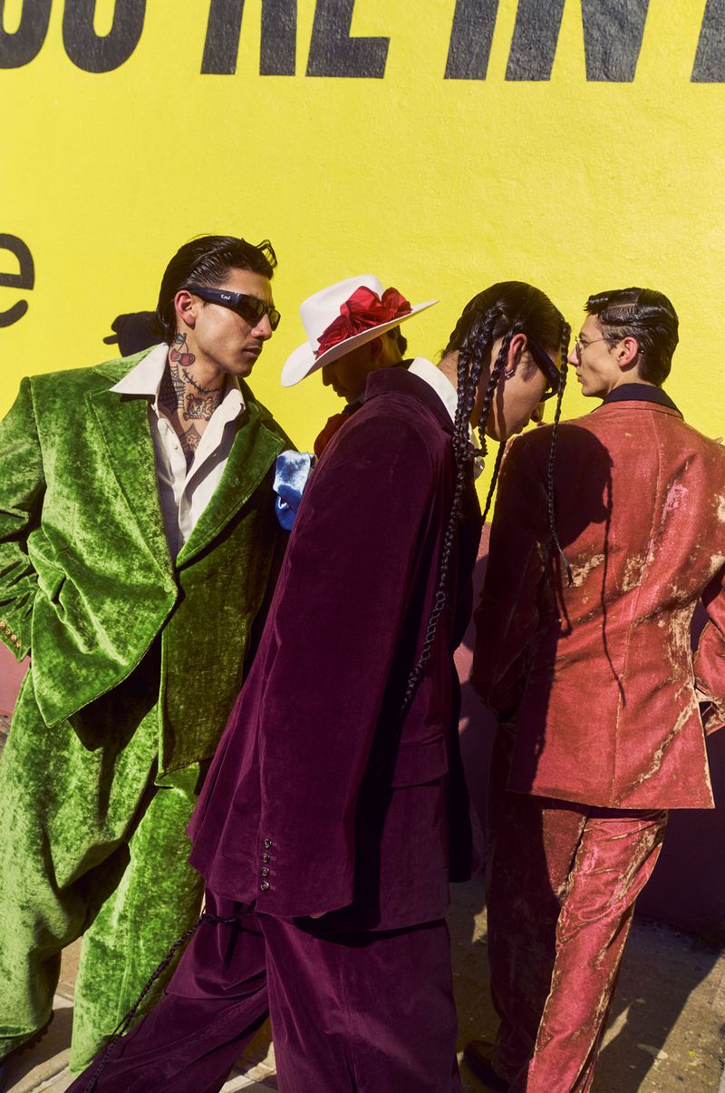 Four people in colorful velvet suits pose against a yellow wall. Fashion shoot shows vibrant colors and textures of velvet fabric.