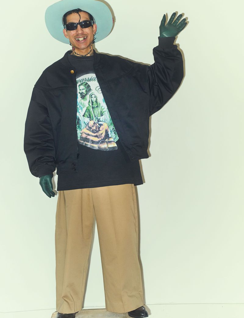 A man in an oversized black jacket, khaki pants, and mint green hat poses against a white wall, wearing sunglasses and gloves.