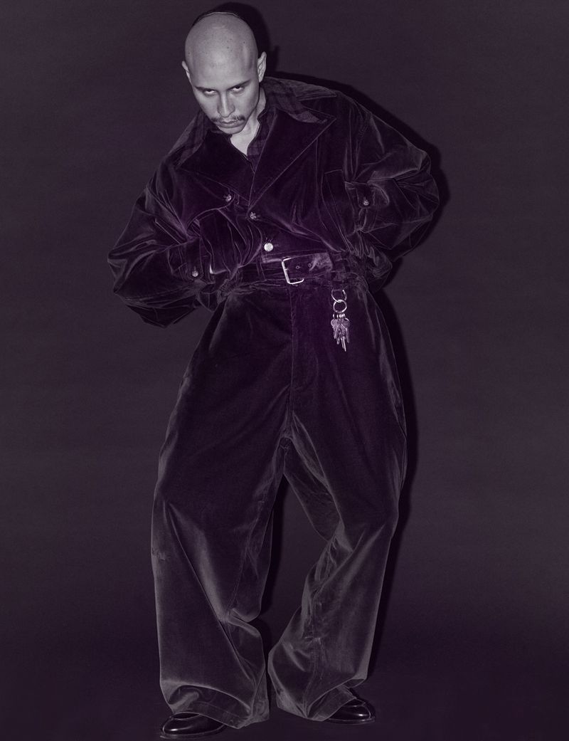 Full-length shot of a bald adult male in a purple velvet suit with a belt, standing in a dark gray photo studio. Fashion editorial style.
