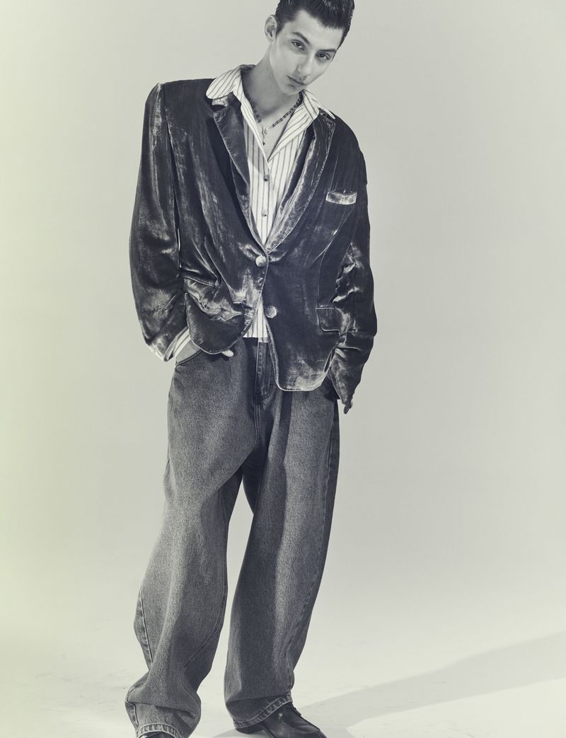 A young male model poses indoors in a velvet jacket, striped shirt, and baggy jeans, highlighted by a neutral background and studio lighting.