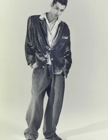 A young male model poses indoors in a velvet jacket, striped shirt, and baggy jeans, highlighted by a neutral background and studio lighting.