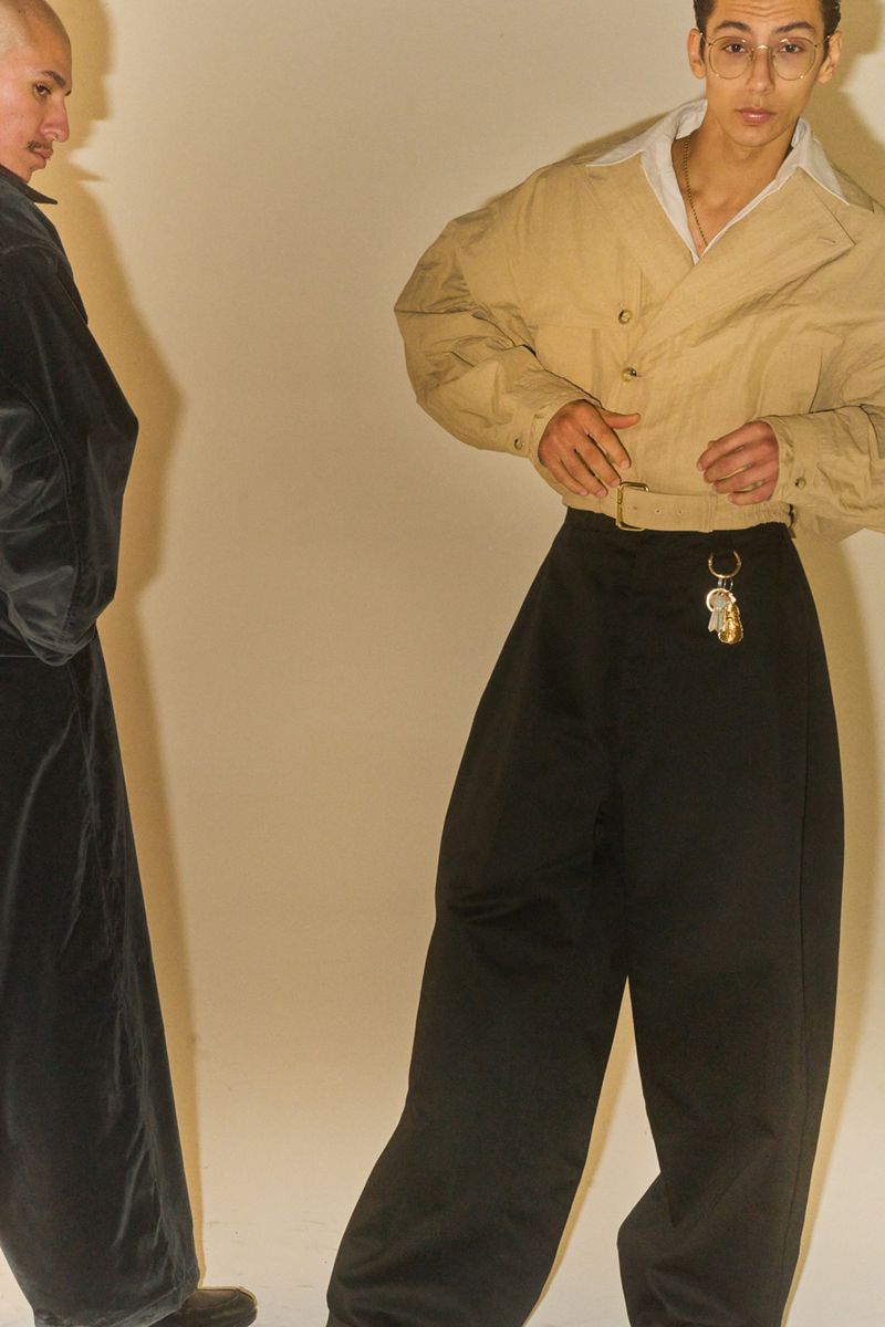 Two adults in a fashion shot, one in black and the other in beige jacket and baggy black pants, in front of a cream-colored backdrop.