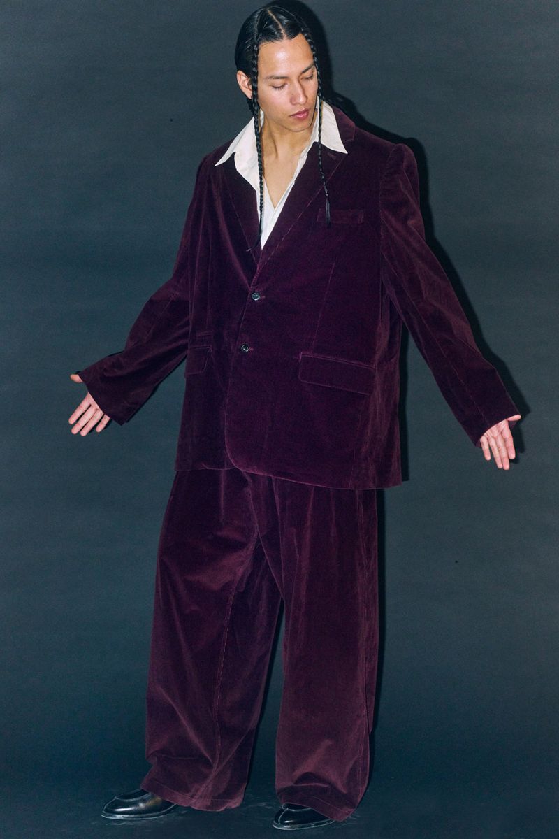 A slender person poses in a maroon velvet suit against a dark background, striking a casual pose with arms slightly outstretched.