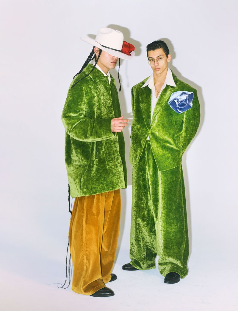 Two stylish adult male models pose in wide-leg pants and matching green velvet suits, set against a clean white backdrop in a fashion editorial.