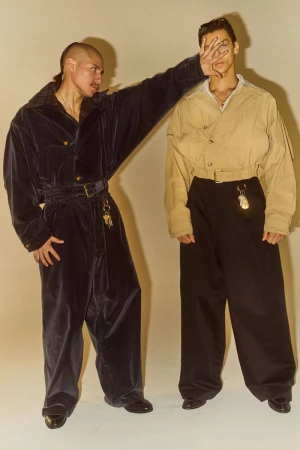Two adult men in baggy jumpsuits pose in a photo studio. One man covers the other's face. Neutral background, eye-level angle.