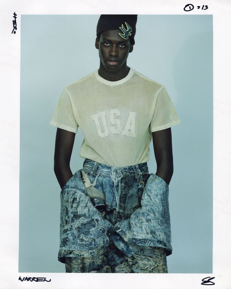 Young adult posing in a t-shirt with USA text, denim jacket wrapped around waist, in a brightly lit studio setting.