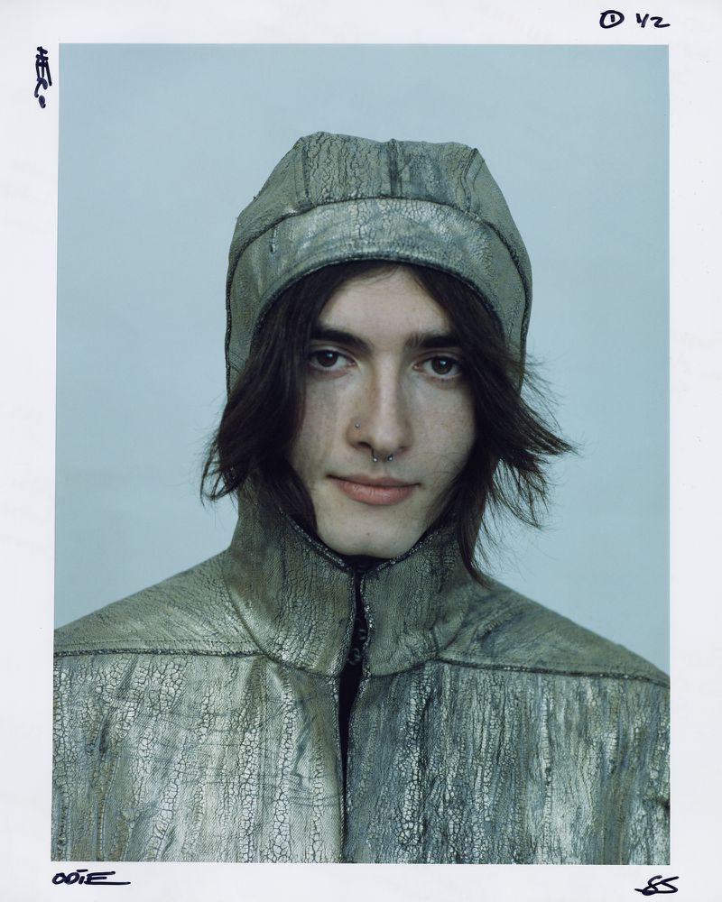 Portrait of a young adult with long brown hair, a nose ring, and a silver metallic hooded jacket in a close-up studio shot.
