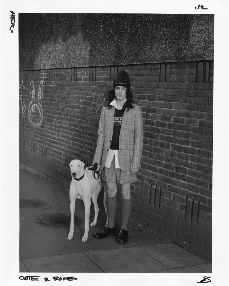 Man and white dog pose against a brick wall in a black and white photograph. The man is wearing a blazer and beanie.