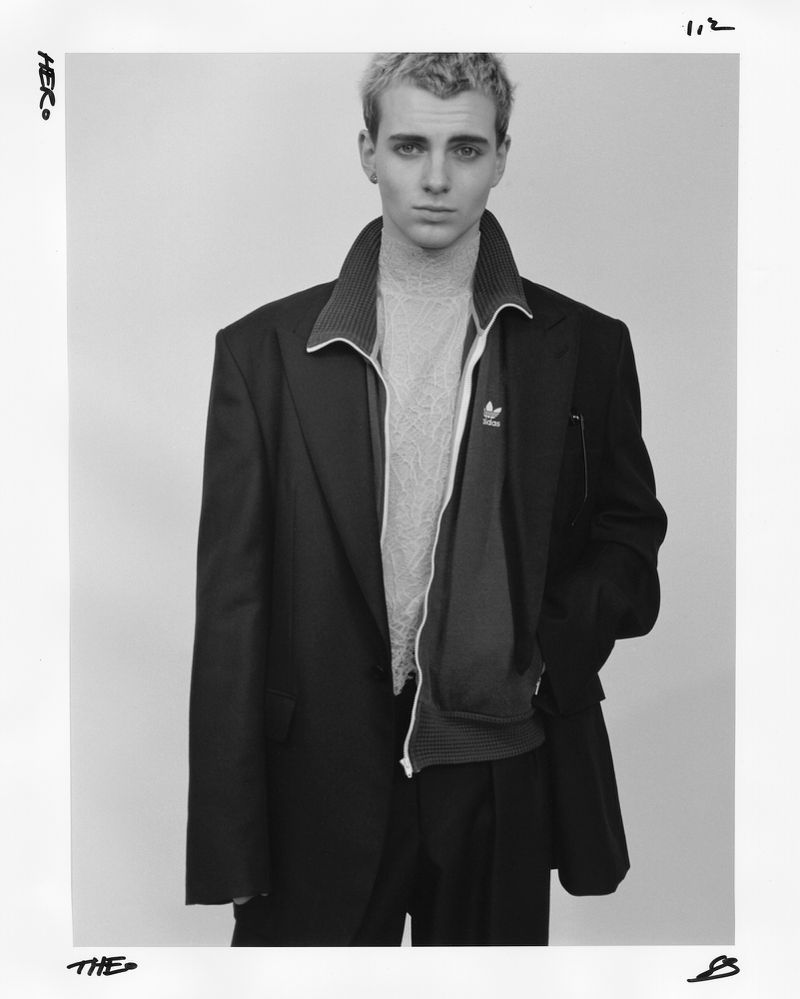 Black and white portrait of a young adult with blond hair, wearing a blazer, sweatshirt, and turtleneck against a grey backdrop.