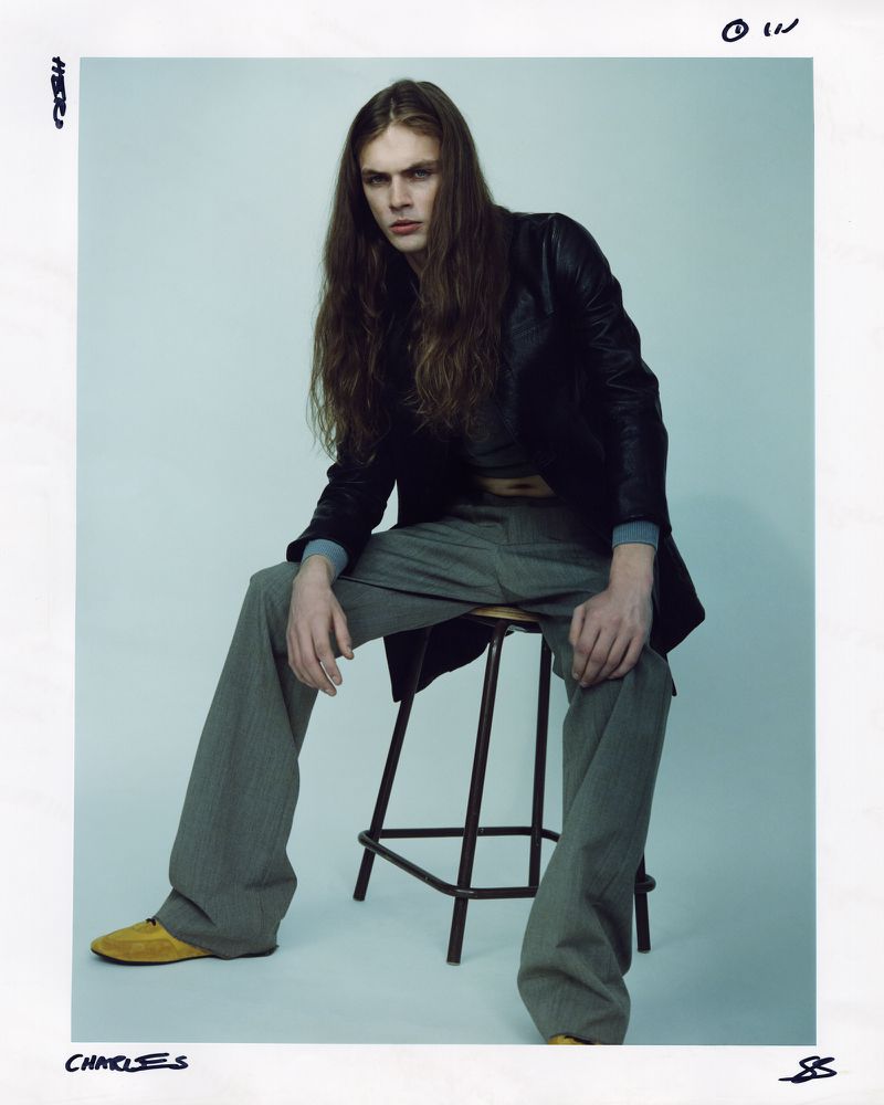 Male fashion model with long hair poses while seated on a stool, wearing a black jacket, grey pants, and yellow shoes in a studio setting.