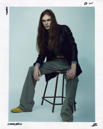 Male fashion model with long hair poses while seated on a stool, wearing a black jacket, grey pants, and yellow shoes in a studio setting.