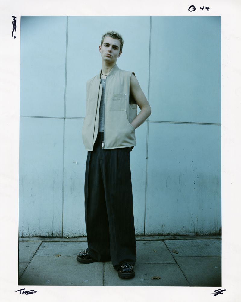 Full-length portrait of a young adult man in a beige vest and dark wide-legged pants against a pale blue architectural wall on a sidewalk.